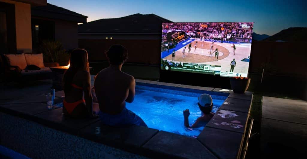 Three people in a pool watching basketball on a large screen installed by the pool.