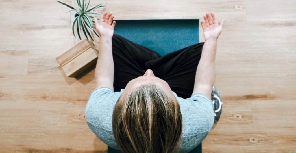 Overhead shot of a person sitting cross-legged on a blue yoga mat with their hands raised in a meditative pose.