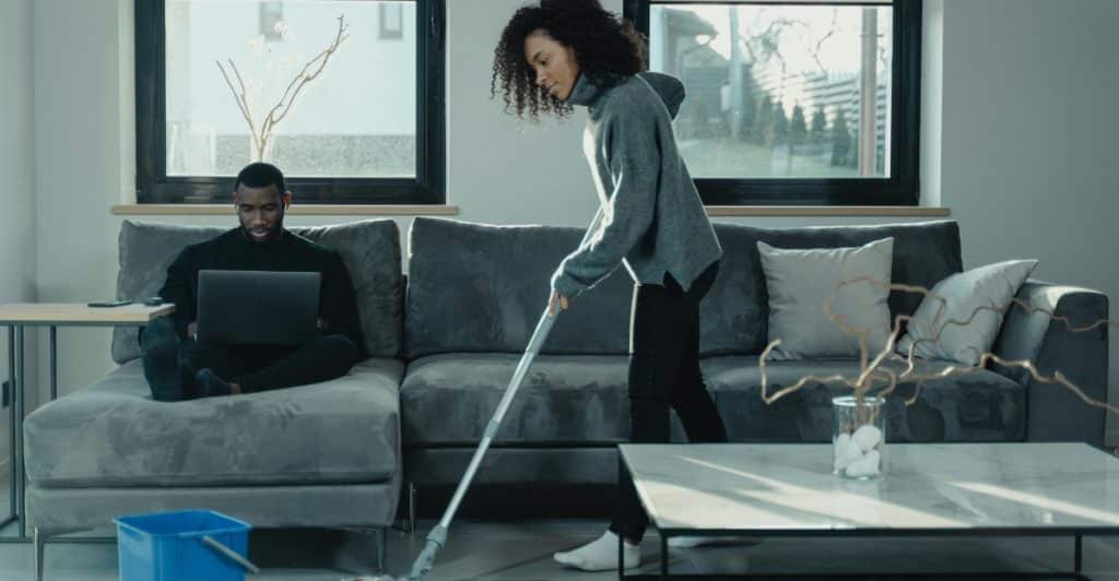 A man using a laptop on a couch while a woman mops the floor in the background, depicting the cleaning of a living or workspace.