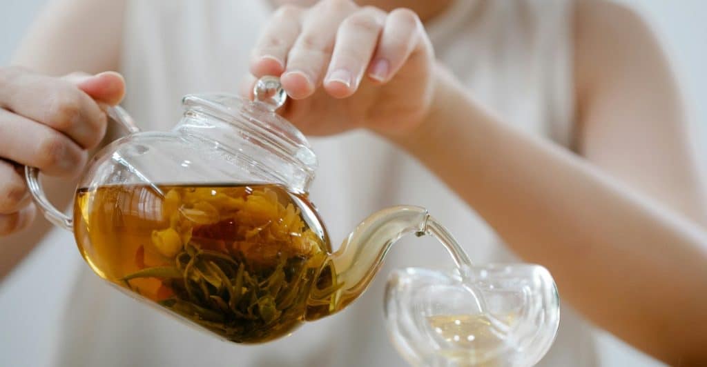 Woman pouring golden-brown tea from a clear glass teapot with tea leaves into a clear glass cup.