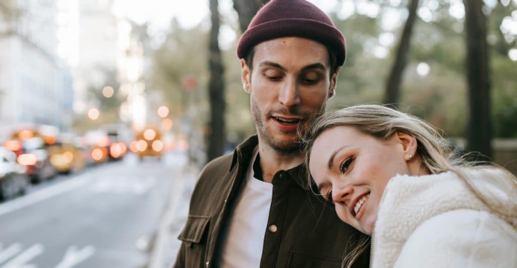 A Man and a woman standing in a street wth the woman resting her head on the man's shoulder.