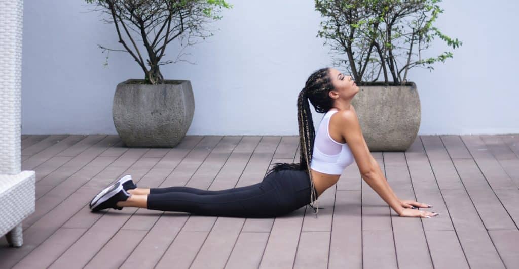 A woman with long dark braids, wearing a white top and dark leggings, stretching in a backbend position in an outdoor patio setting with potted plants.