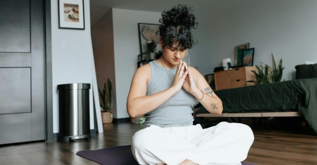 A woman with curly hair sitting cross-legged on a yoga mat with her hands in a prayer position, meditating in a room.
