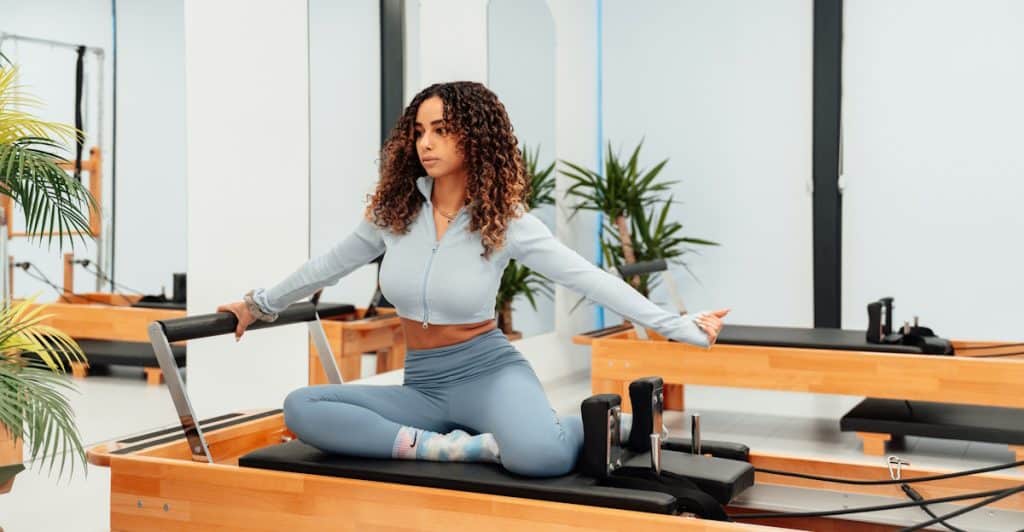 A woman with curly brown hair, wearing light blue athletic wear, sitting on a Pilates reformer machine in a bright studio with mirrors and plants.