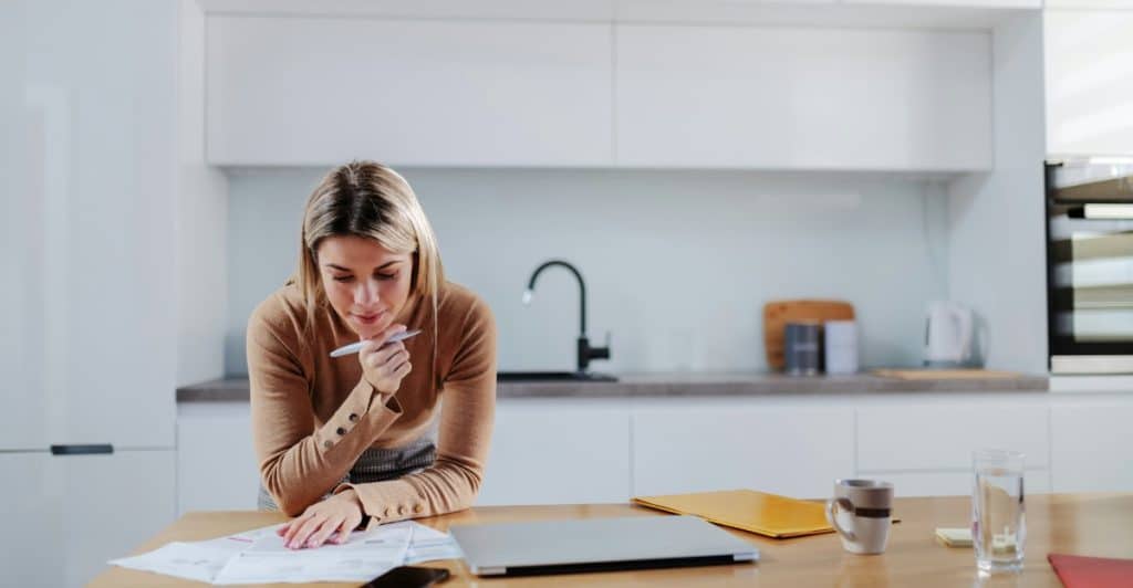 A blonde woman in a brown sweater reviewing papers at a kitchen table with a laptop and other items.