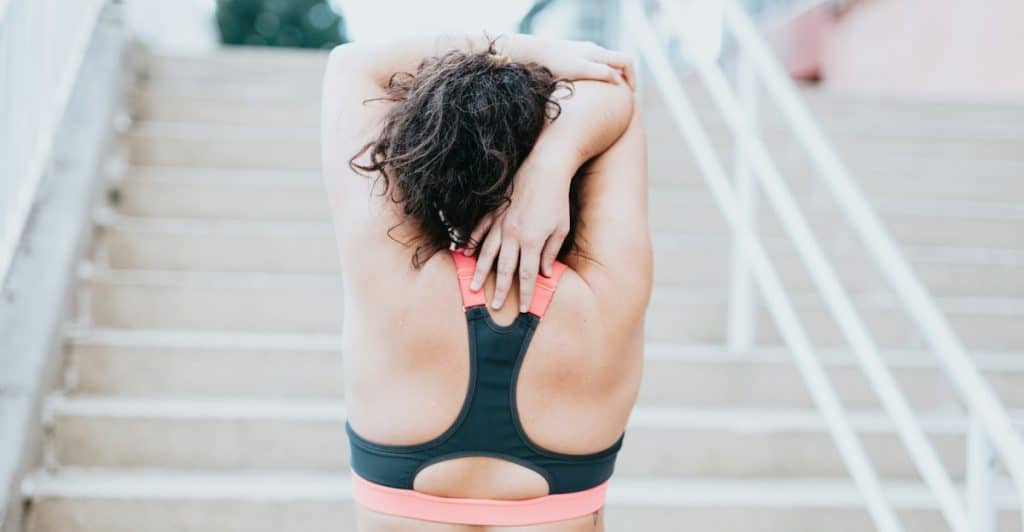 A woman in athletic wear stretching her arm behind her back while standing on outdoor steps