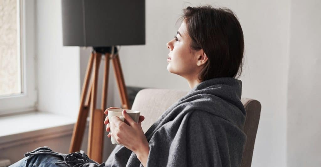 A woman wrapped in a blanket sitting in a chair, holding a mug and looking out a window.