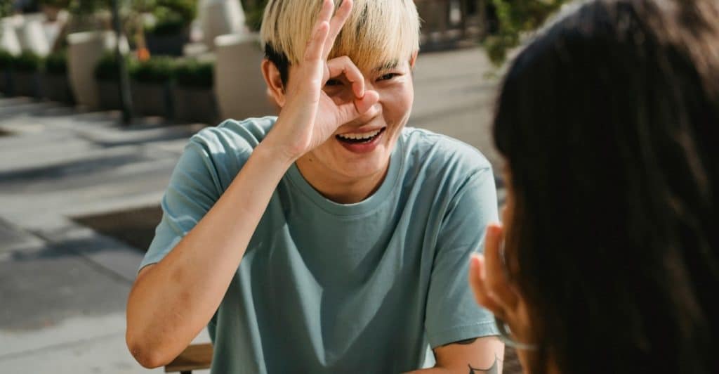  A young person with dyed blonde hair and tattoos, wearing sunglasses, makes an "okay" hand gesture near their eye while sitting at an outdoor table.