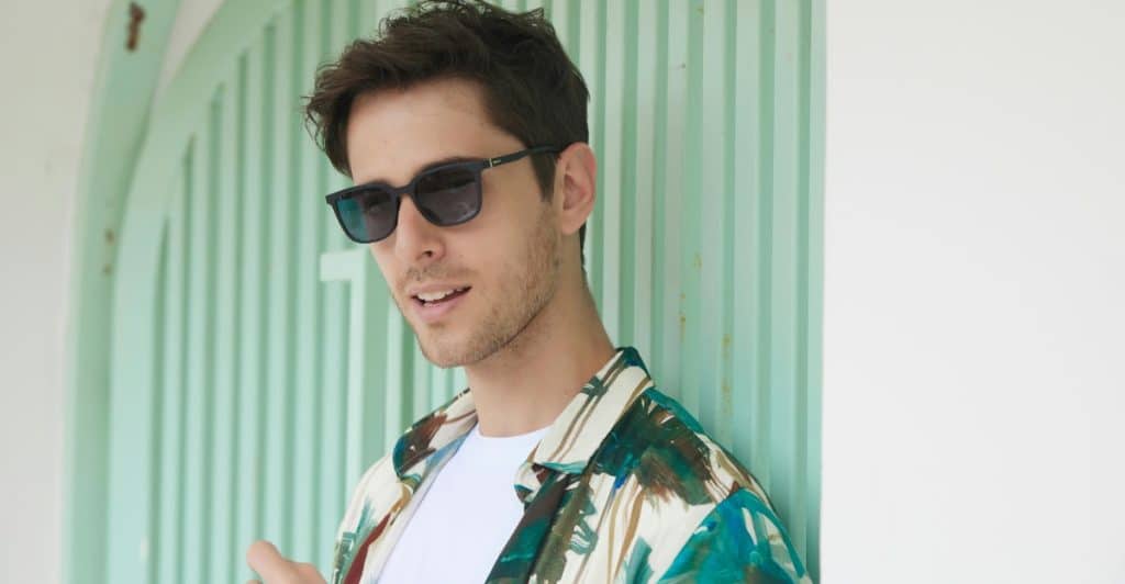 A man wearing sunglasses and a patterned shirt, leaning against a light green wall.