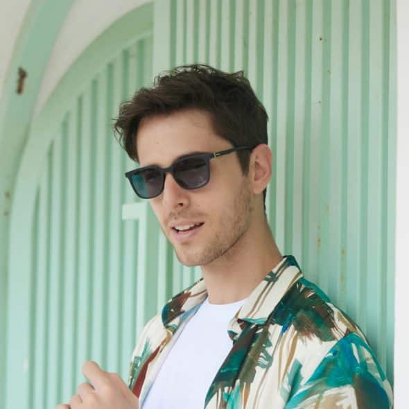 A man wearing sunglasses and a patterned shirt, leaning against a light green wall.