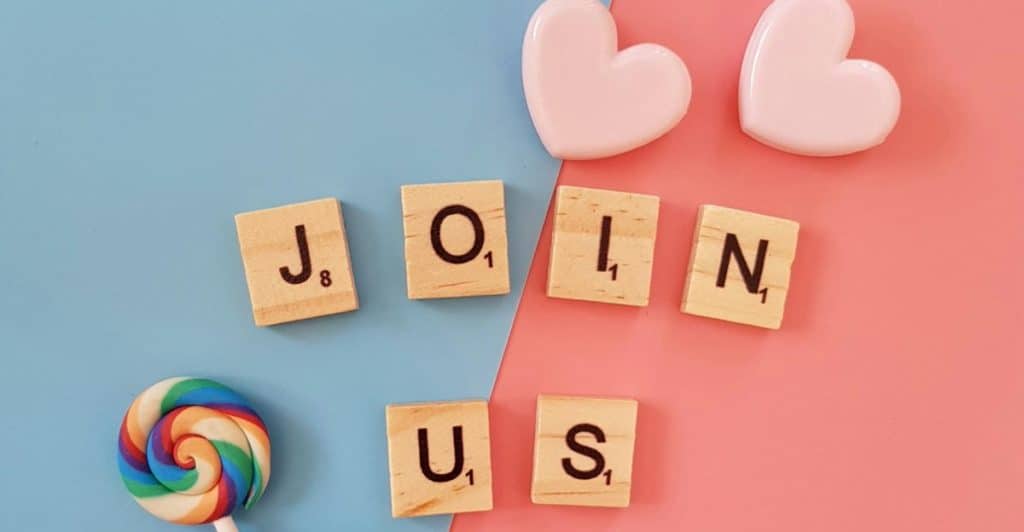 Scrabble tiles spelling "Join Us" are arranged on a split pink and blue background, along with heart decorations and lollipops.
