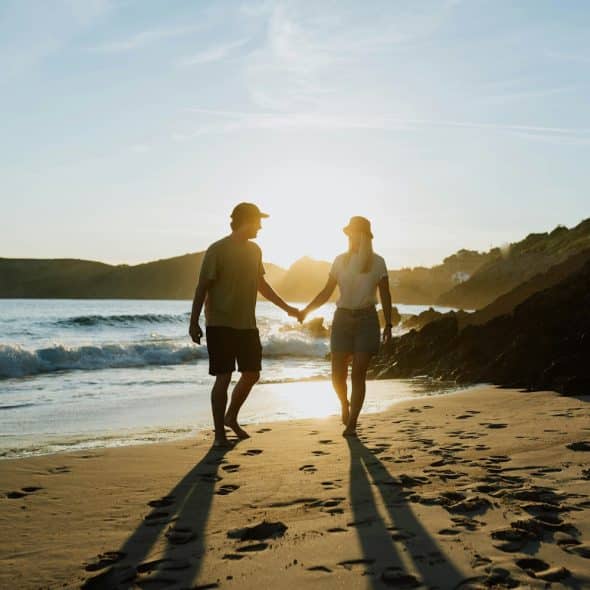 A couple holding hands and walking barefoot on a sandy beach towards the ocean as the sun sets, casting long shadows behind them.