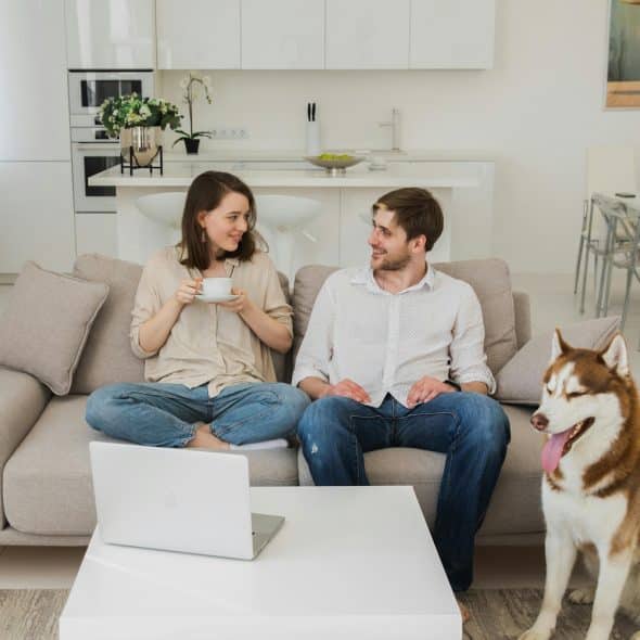 A man and a woman are sitting on a sofa in a living room with a husky dog sitting beside them.