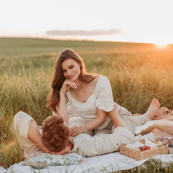 A man lying on a picnic blanket in a field with a woman sitting on his lap, both smiling at the camera.
