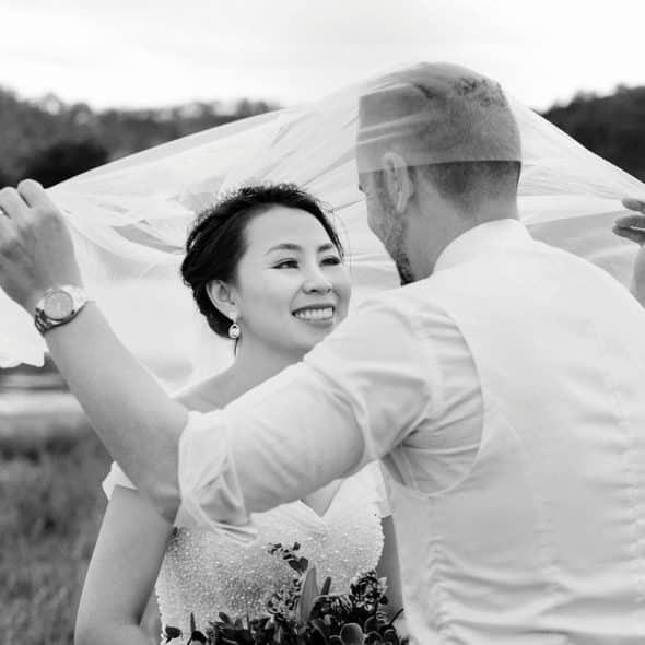 A groom lifting the veil of his smiling bride in a black and white outdoor wedding photo.
