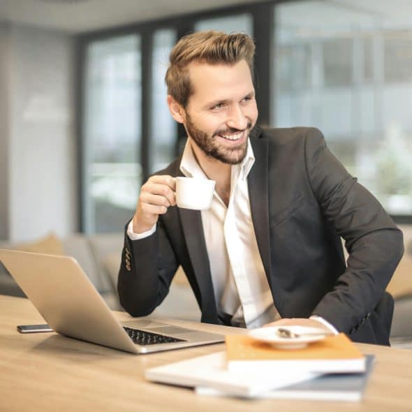 A smiling man in a suit jacket and open-collared shirt is seated at a light wooden desk, holding a white coffee cup to his mouth with his right hand, and looking towards the right while a silver laptop and some books are on the desk in front of him.