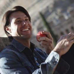A man with dark hair smiling brightly while holding a red apple, sitting outdoors with an old stone building in the background.