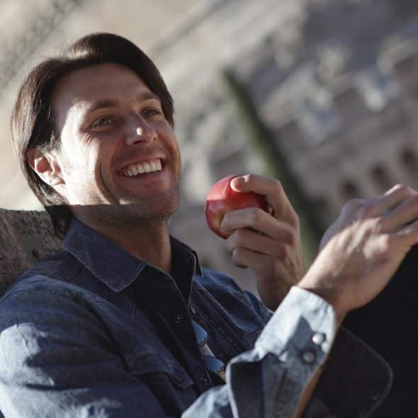 A man with dark hair smiling brightly while holding a red apple, sitting outdoors with an old stone building in the background.