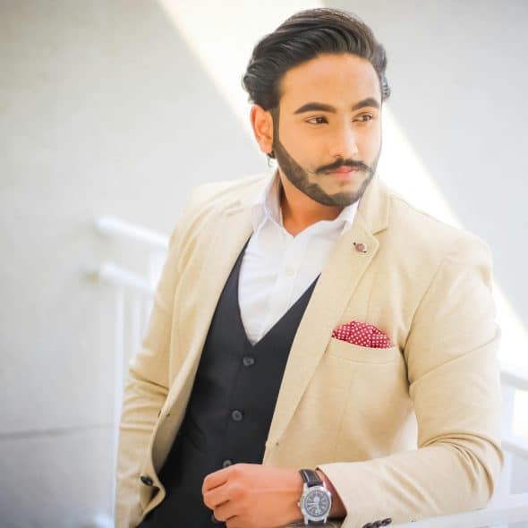 A man in a beige suit, black vest, and white shirt with a red and white polka dot pocket square, posing with one hand in his pocket and the other arm resting on a railing, wearing a watch.