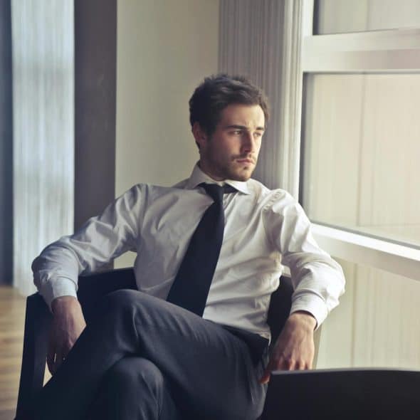 A man in a white shirt and black tie looking out a window while sitting on a chair.