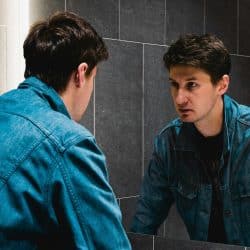 A man in a denim jacket looking intently at his reflection in a mirror.