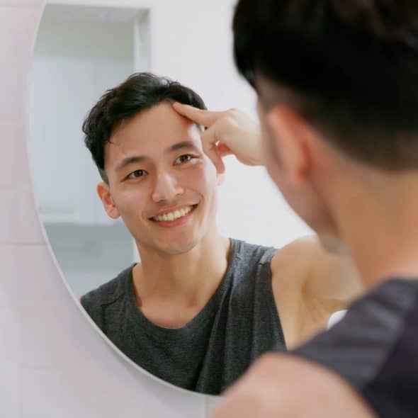 A man looking in a mirror and smiling while touching his forehead.