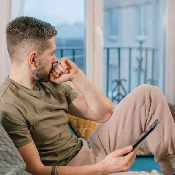 A man in a green shirt and beige pants sitting on a couch, looking out a window with a pensive expression, holding a tablet in his right hand.