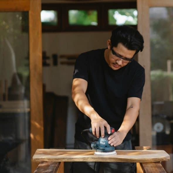 A man sanding a wooden plank with a power sander, wearing safety glasses.