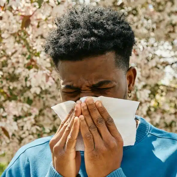 A man who appears to be Black or African American sneezing into a tissue in front of a blossoming tree.