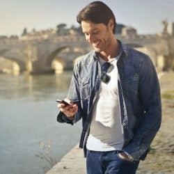A man standing by a river, looking down and smiling at his phone with a bridge in the background.
