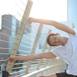 A man in a white t-shirt and black shorts stretching his leg on a railing with city buildings in the background.