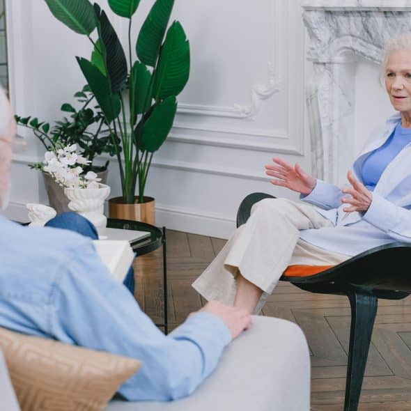 A woman with gray hair wearing a light blue top and white shirt talking animatedly to a man while sitting on a patterned chair in a well-decorated living room.