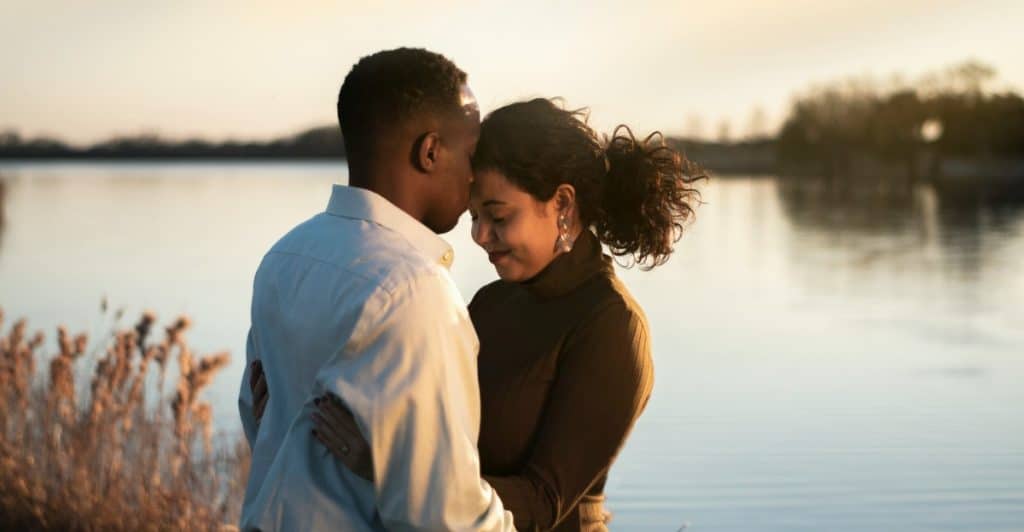 A man and a woman embracing by a lake, with the man kissing the woman's forehead.
