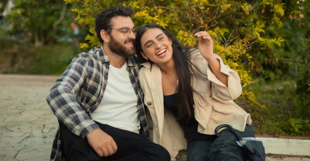 A man with glasses and a plaid shirt sitting next to a woman with long dark hair, both laughing outdoors.
