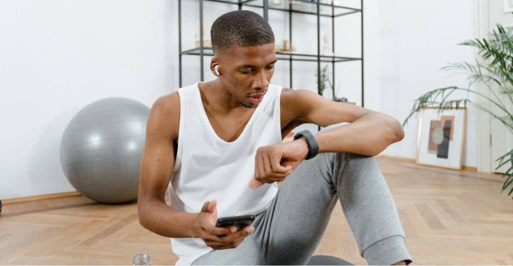 A lean, black man in the gym is looking at his stopwatch and holding a mobile while sitting on the floor.