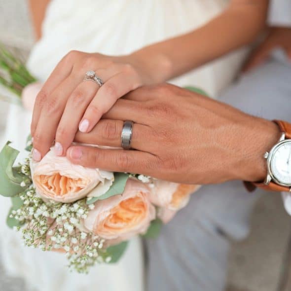 A newlywed couple's hands are shown, with the woman's hand gently resting on top of the man's, displaying their wedding rings, and a bouquet of peach-colored flowers is visible in the foreground.