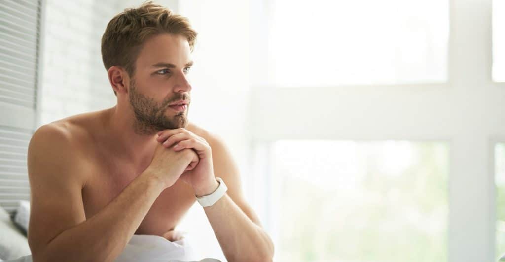A shirtless man with a beard sitting on the edge of a bed with his hands clasped looking towards a brightly lit window.