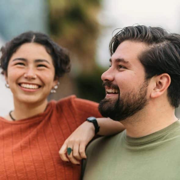 A smiling woman with short, dark curly hair and a man with a beard, both looking to the side and smiling.
