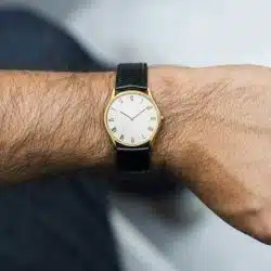 A man's forearm with a gold-tone watch with a white dial and black leather strap, positioned to show the watch clearly on the wrist.