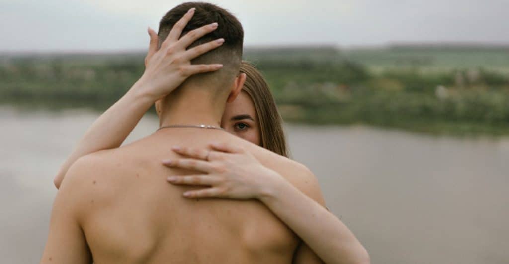 A woman with long hair hugging a shirtless man with a lake in the background