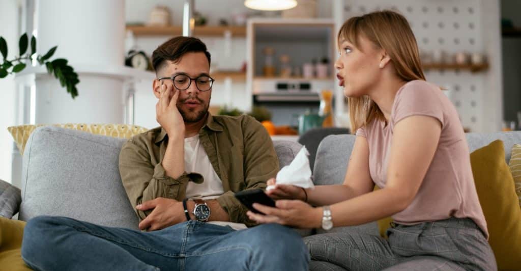 A woman sitting on a couch holding a phone and gesturing towards a man who is looking at the phone with a surprised expression.