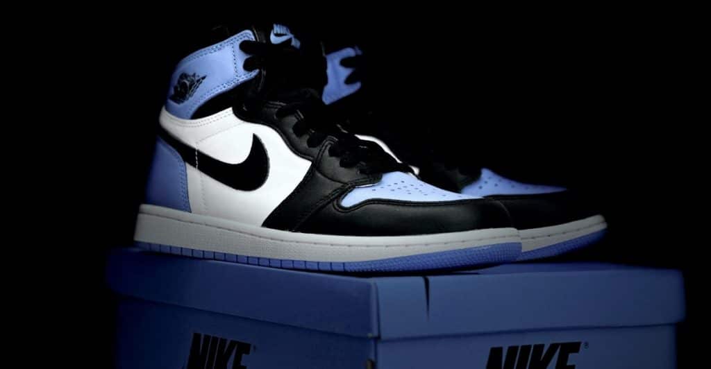 A black, white, and light blue Air Jordan 1 Retro High OG sneaker sitting on a blue Nike shoe box against a black background.