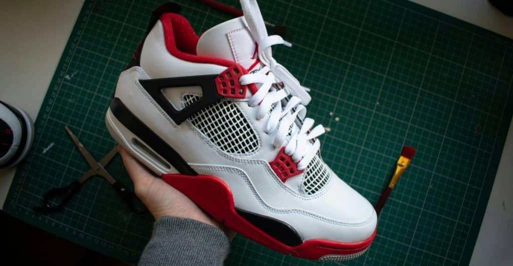 A person's hand holding a white, black, and red Air Jordan 4 sneaker with visible mesh and plastic wings, set against a green mat with scissors and paper clips.