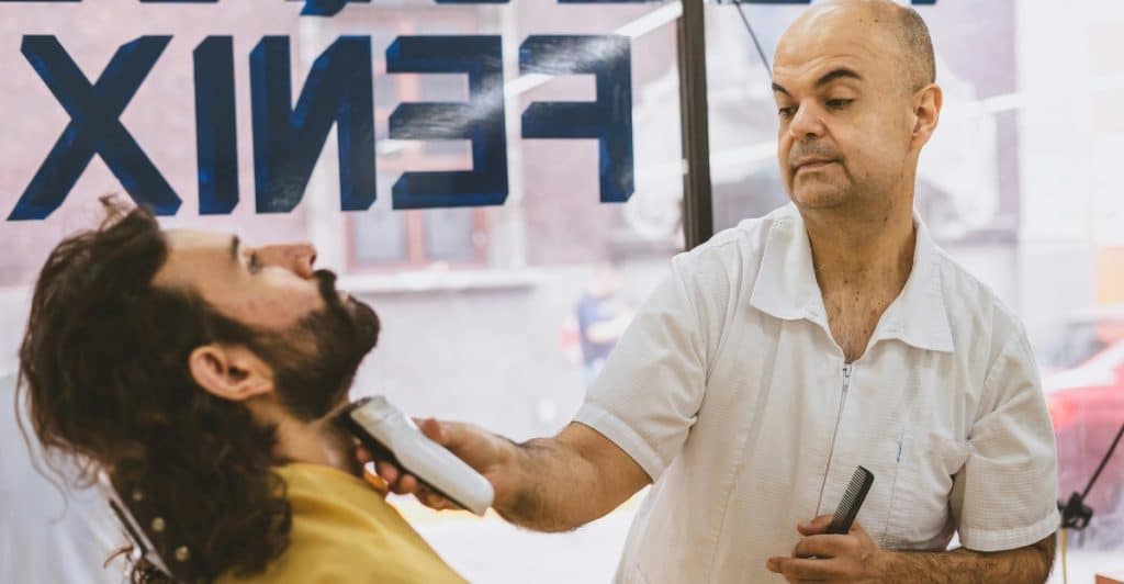 A barber trimming a man's beard with an electric razor, holding a comb in his other hand.