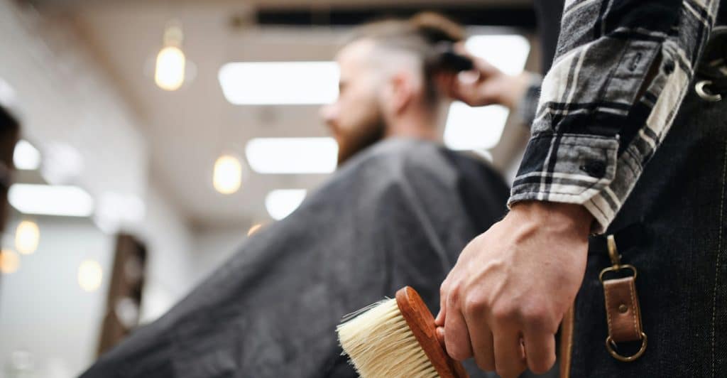 A close-up of a barber's hand holding a wooden neck duster brush with a client getting a haircut in the background.