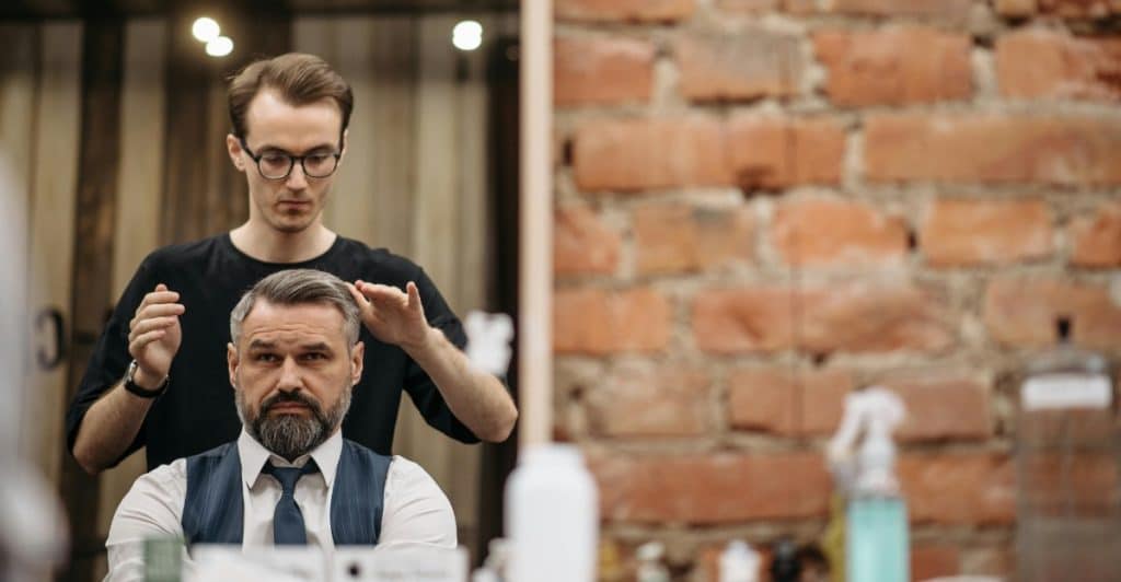 A barber with glasses styling a man's short, textured hair in front of a mirror.