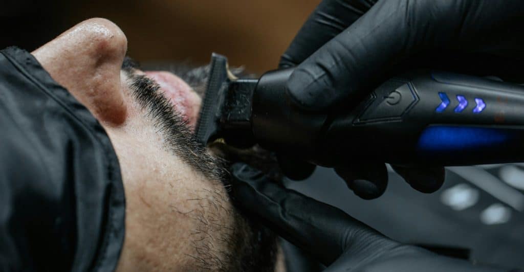 A close-up of a barber's gloved hands using an electric trimmer on a client's beard.