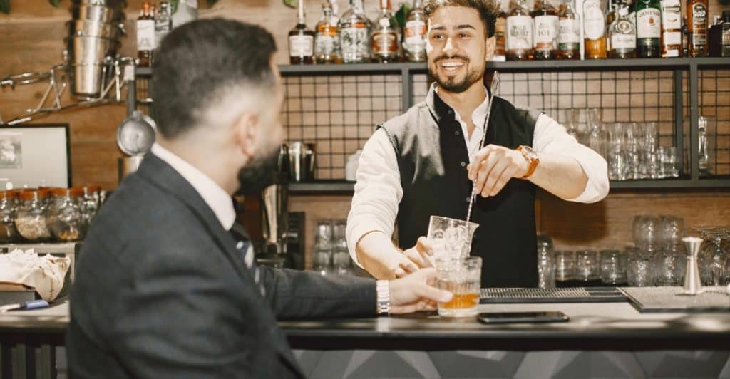 A smiling bartender with a beard and a vest mixing a drink, looking at a customer seated at the bar who is holding a glass of whiskey.