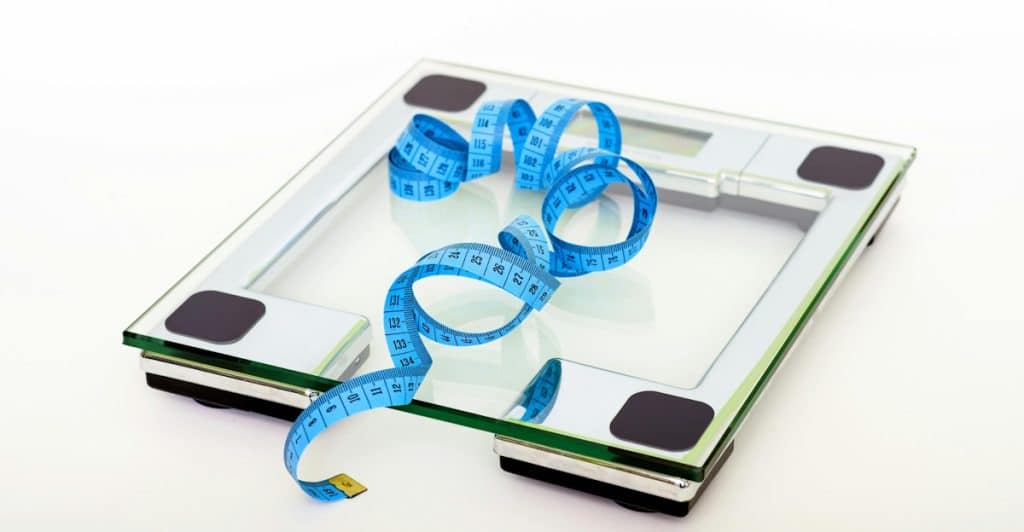 A clear glass digital bathroom scale with a blue measuring tape wrapped around it, on a white surface.