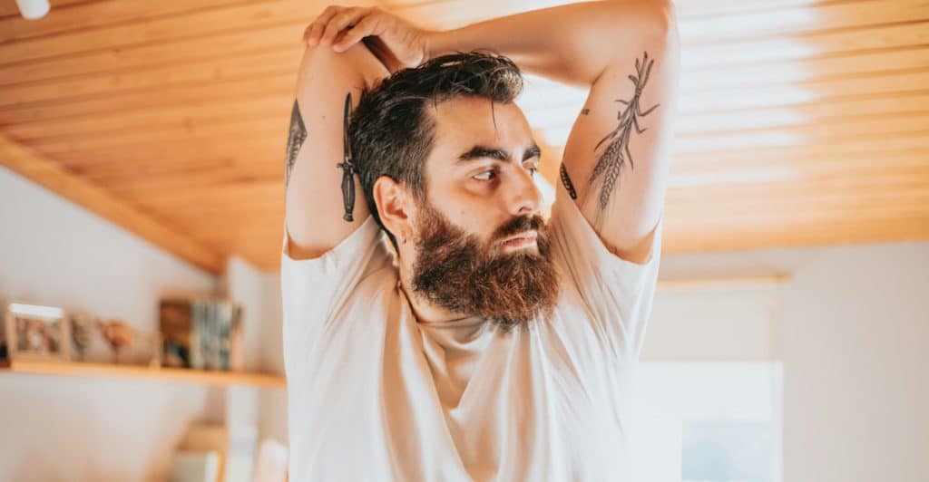 A man with a full beard and arm tattoos stretching his arms behind his head in a sunlit room.